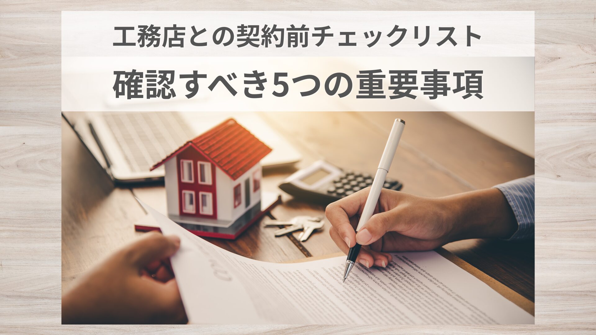契約書に署名する手元と、模型の家・電卓・鍵が置かれた木製デスクの上の様子